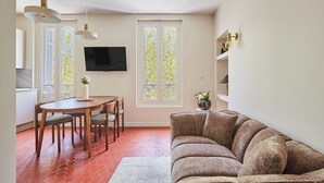 Un salon comprenant un canapé, une table à manger avec des chaises, un téléviseur fixé au mur et une fenêtre donnant sur des arbres.
