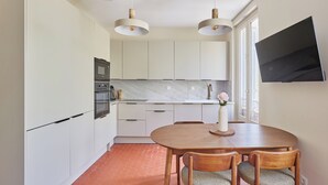 Une cuisine moderne avec des meubles blancs, une table à manger avec des chaises, un téléviseur fixé au mur et des luminaires suspendus.
