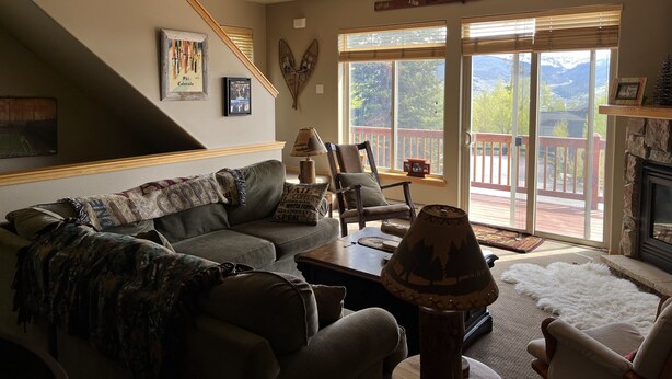 Ein Wohnzimmer mit einer Couch, einem Sessel, einer Lampe und einem Kamin. Eine Treppe führt zu einer anderen Ebene, und ein großes Fenster bietet Blick auf die Berge.
