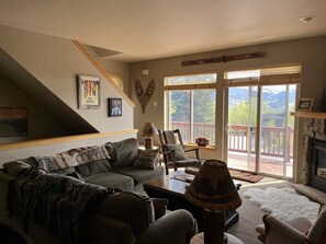 Un salon comprenant un canapé, un fauteuil, une lampe et une cheminée. Un escalier mène à un autre niveau, et une grande fenêtre offre une vue sur les montagnes.