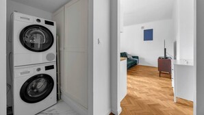 Una lavanderia con lavatrici affiancate e un corridoio che conduce a un soggiorno con un divano e un mobile TV.