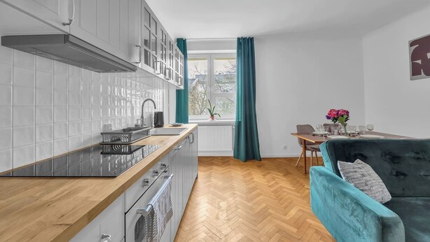 Una cucina moderna con mobili bianchi, un piano di lavoro in legno e un'area pranzo con un divano color petrolio e un tavolo apparecchiato per quattro persone.
