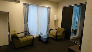 Ein Hotelzimmer mit zwei Sesseln, einem Glastisch, einem großen Fenster mit Vorhängen, einer Tür und einem sichtbaren Badezimmer.