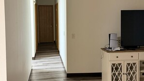 Un pasillo con suelo de madera, una pared blanca, un televisor sobre un mueble y una puerta al final.