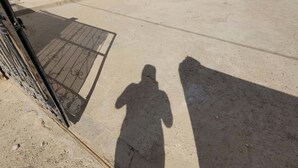 Une cour en béton avec un portail en métal, un bâtiment blanc aux fenêtres grillagées et l’ombre d’une personne.