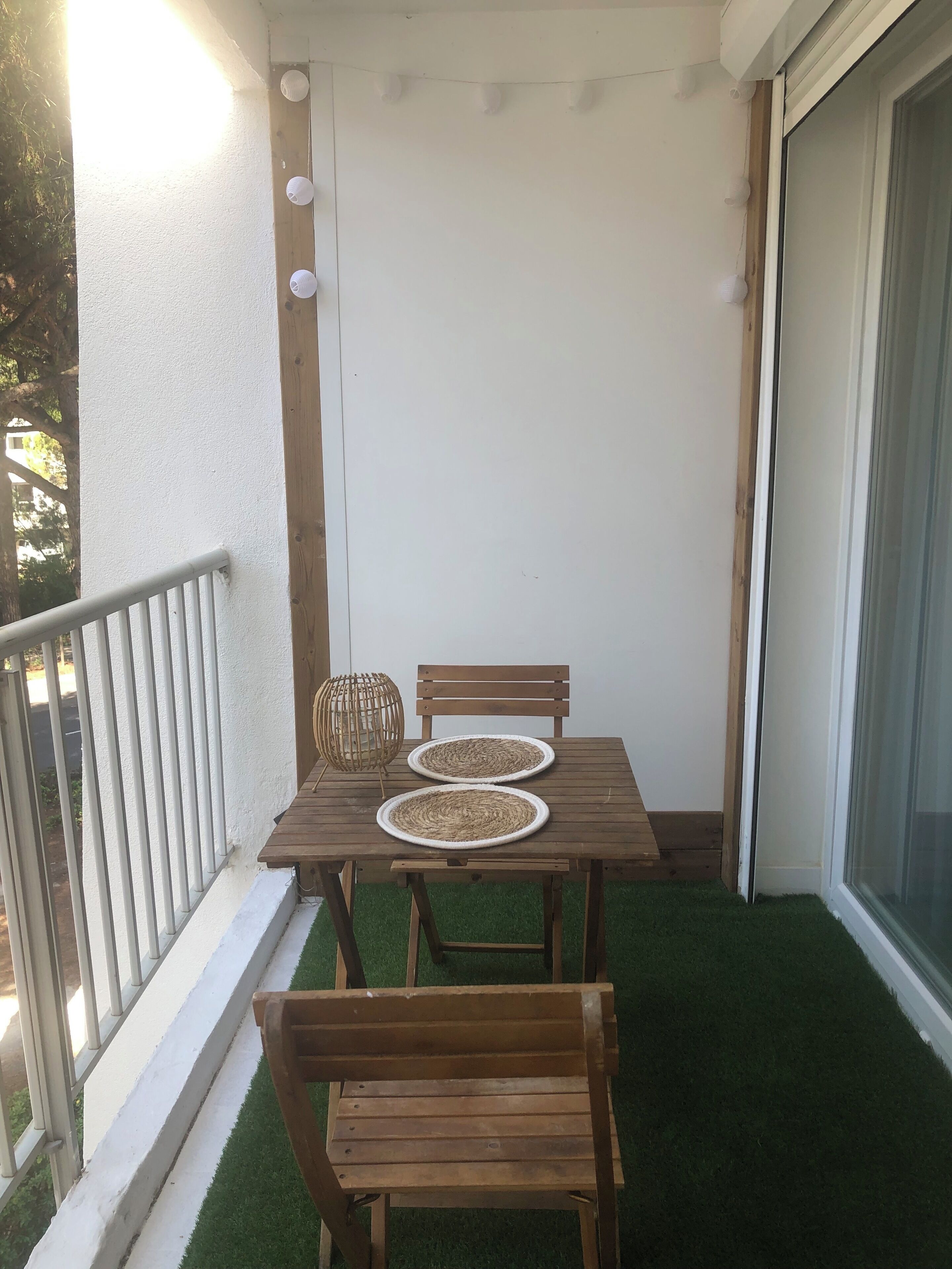Un balcón con una mesa y sillas de madera, una cesta tejida y una pared blanca con una ventana.
