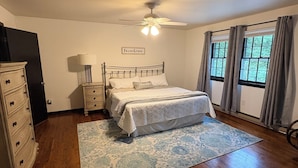 Une chambre à coucher comprenant un lit, une commode, une table de chevet, une lampe, un ventilateur de plafond et des fenêtres avec des rideaux.