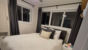 Un dormitorio con cama, mesita de noche, ventilador de techo y ventana con cortinas.