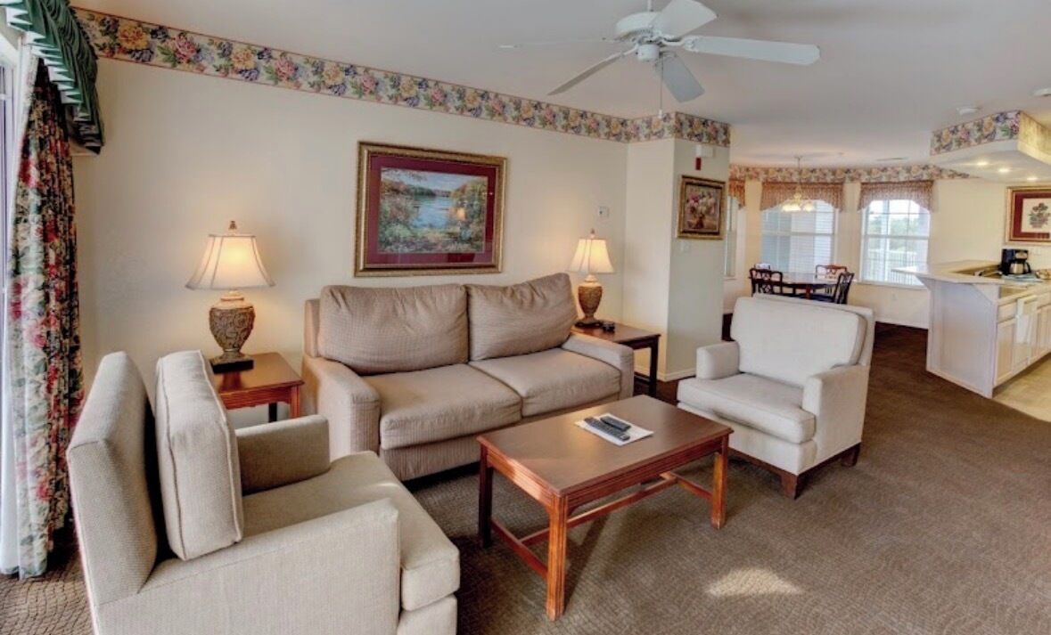 A living room with a sofa, armchairs, a coffee table, and lamps. There is a painting on the wall and a ceiling fan.