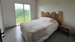 Camera da letto con un letto grande, una testiera in legno, una piccola panca di legno e una porta scorrevole in vetro che dà su un balcone con vista sul verde.