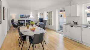 Et moderne køkken med hvide skabe, et spiseområde med et rundt bord og stole, samt en opholdsstue med sofa og fjernsyn.