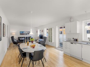 Eine moderne Küche mit weißen Schränken, ein Essbereich mit rundem Tisch und Stühlen sowie ein Wohnbereich mit Sofa und Fernseher.