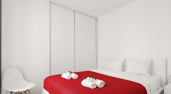 A modern hotel room with a bed, white walls, a red accent wall, a white chair, and toiletries on the bed. - Charmant pied-à-terre parisien au cœur du Marais • 4 pers (Paris)