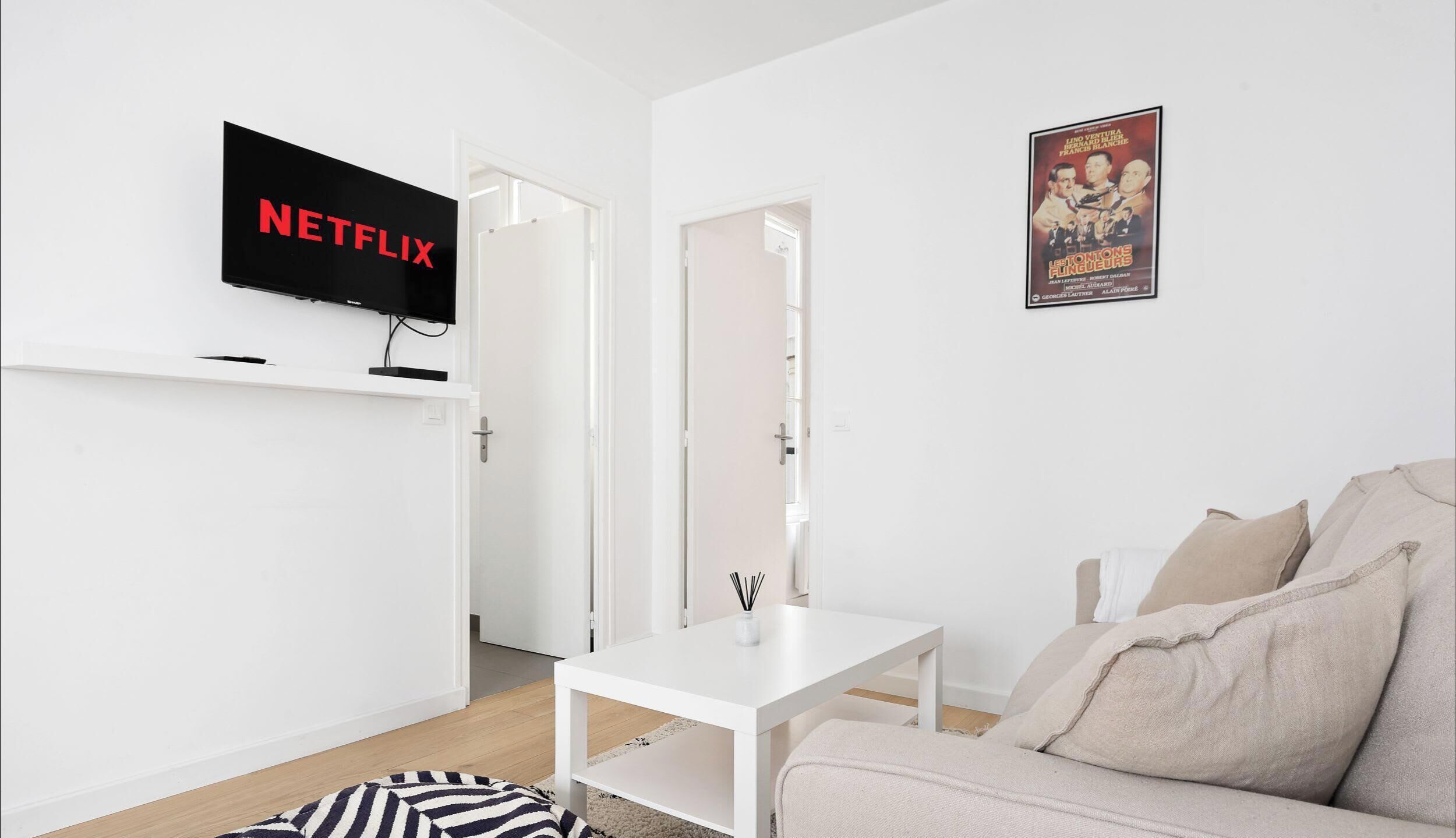 A modern living room with a white sofa, a small white table, a mounted TV displaying 'NETFLIX', and a poster on the wall.