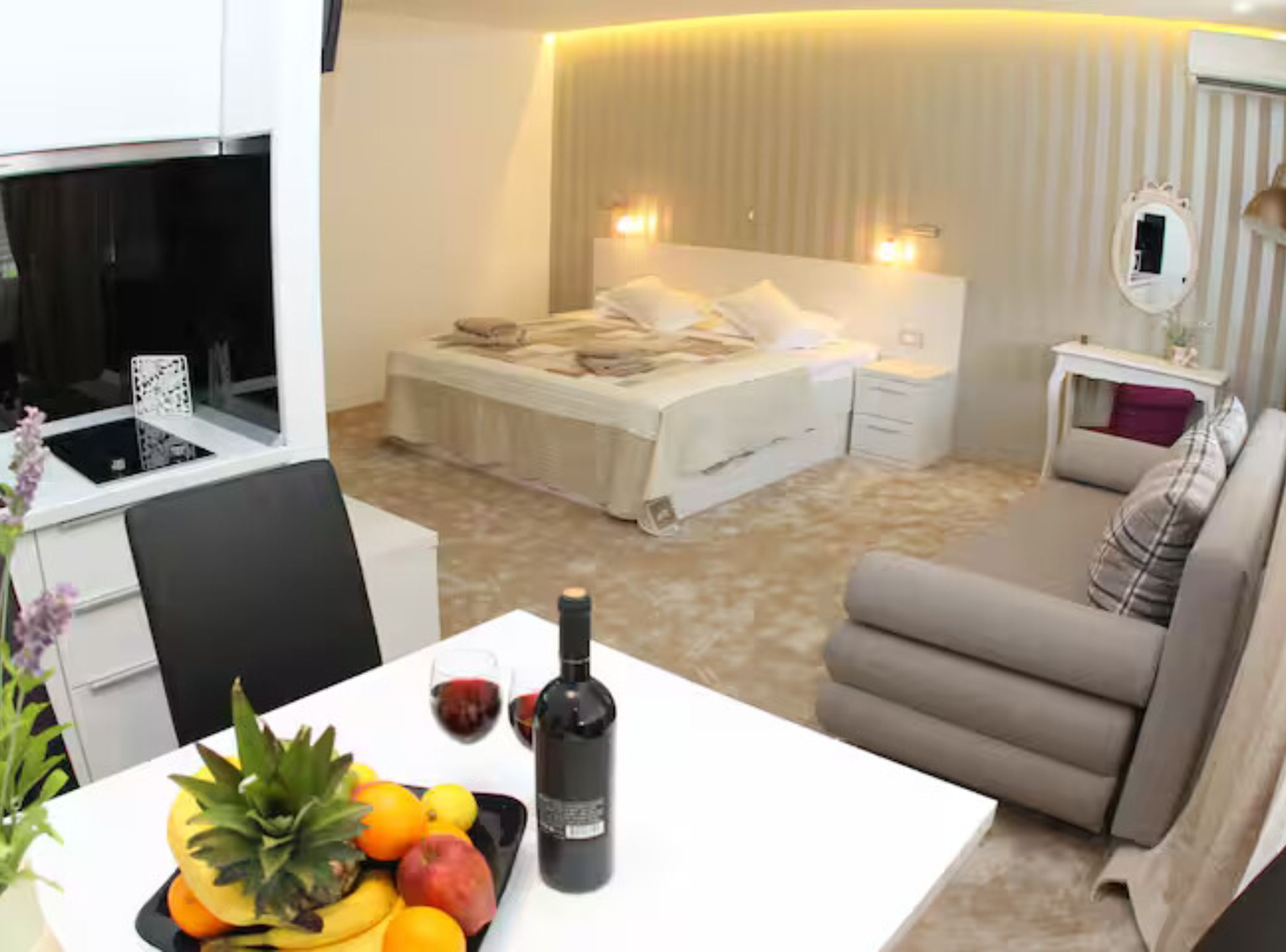 A hotel room with a bed, a sofa, a dining table with fruit and a bottle, and a small table with a lamp.