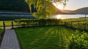 Asfaltoitu polku johtaa nurmialueelle, jossa on aita ja puu. Alueelta avautuu näkymä vesistöön ja kukkuloille.