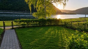 En asfalteret sti fører til et græsområde med hegn og et træ, med udsigt over vand og bakker.