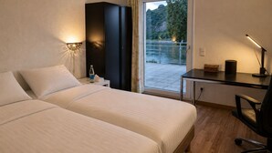 Hotellihuone, jossa on kaksi sänkyä, työpöytä, tuoli, lamppu, yöpöytä, vesipullo ja näkymä joelle.