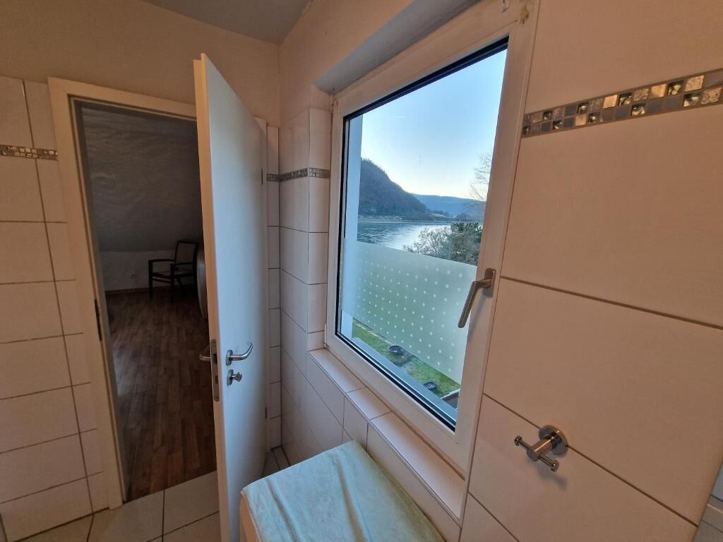 A bathroom with a window view of a river and hills, a door leading to another room, and tiled walls.