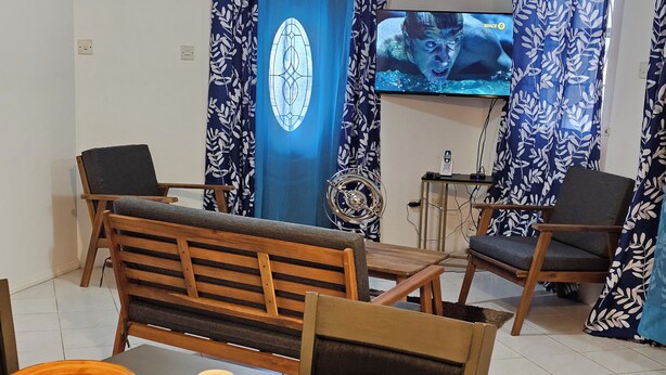 Een eetgedeelte met een houten bank, twee stoelen en een tafel gedekt met borden, bestek en een glazen schaal. Een televisie is aan de muur bevestigd, en er is een gordijn met een patroon te zien.