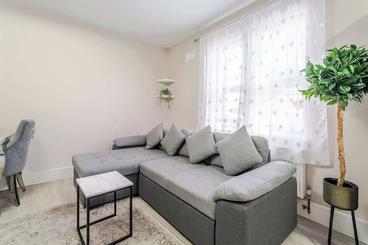 Una sala de estar con un sofá seccional gris, una mesita, una silla, una planta en maceta y una ventana con cortinas de tul.