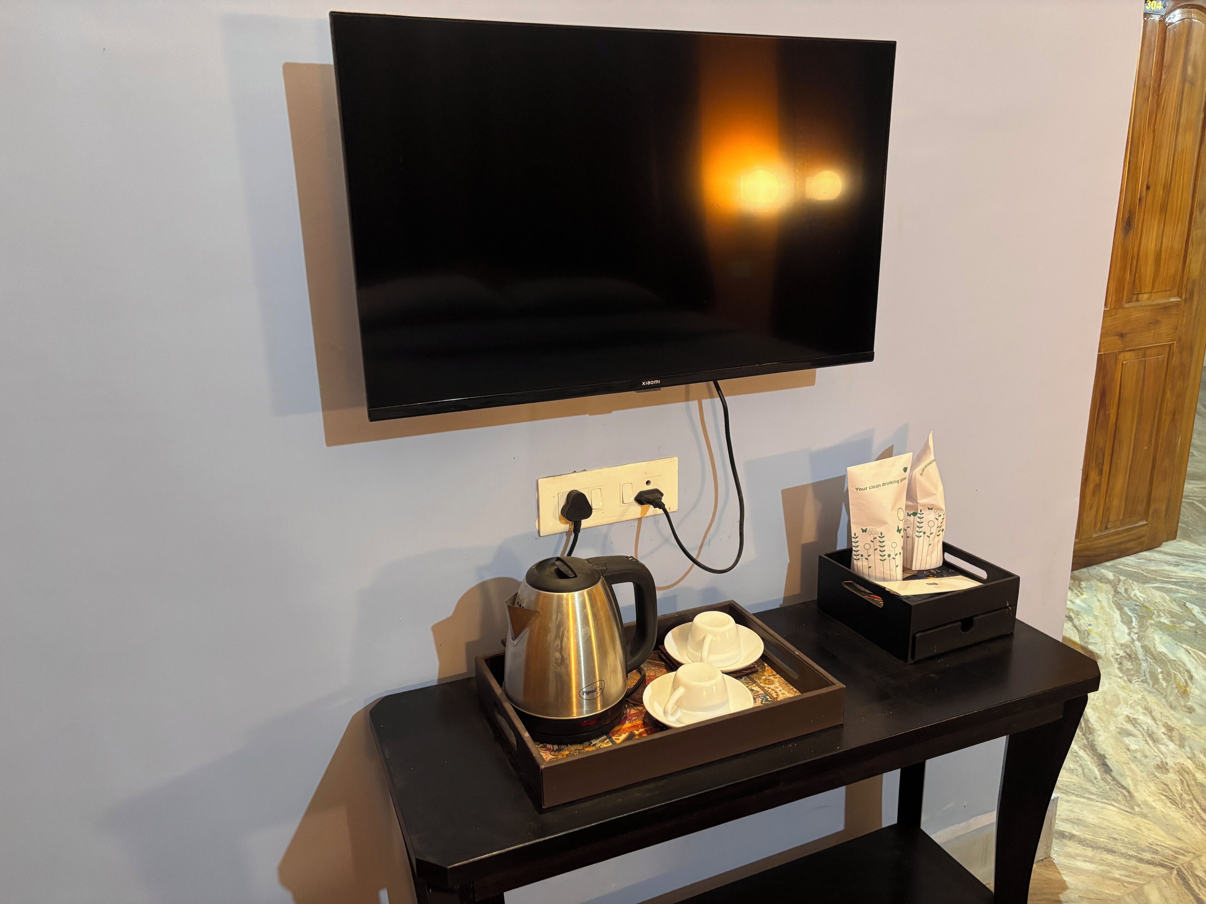 A flat-screen TV mounted on a wall, a coffee maker, and a tray with two cups and a bag of coffee pods on a small table.