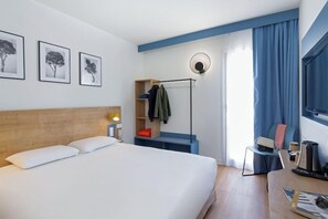 A hotel room with a bed, a desk, a chair, a coat rack, and wall art. - Kyriad Marseille Est - Aubagne Gemenos (Gemenos)