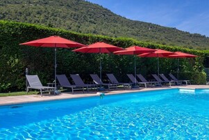 A swimming pool with red umbrellas, lounge chairs, and a hedge in the background.