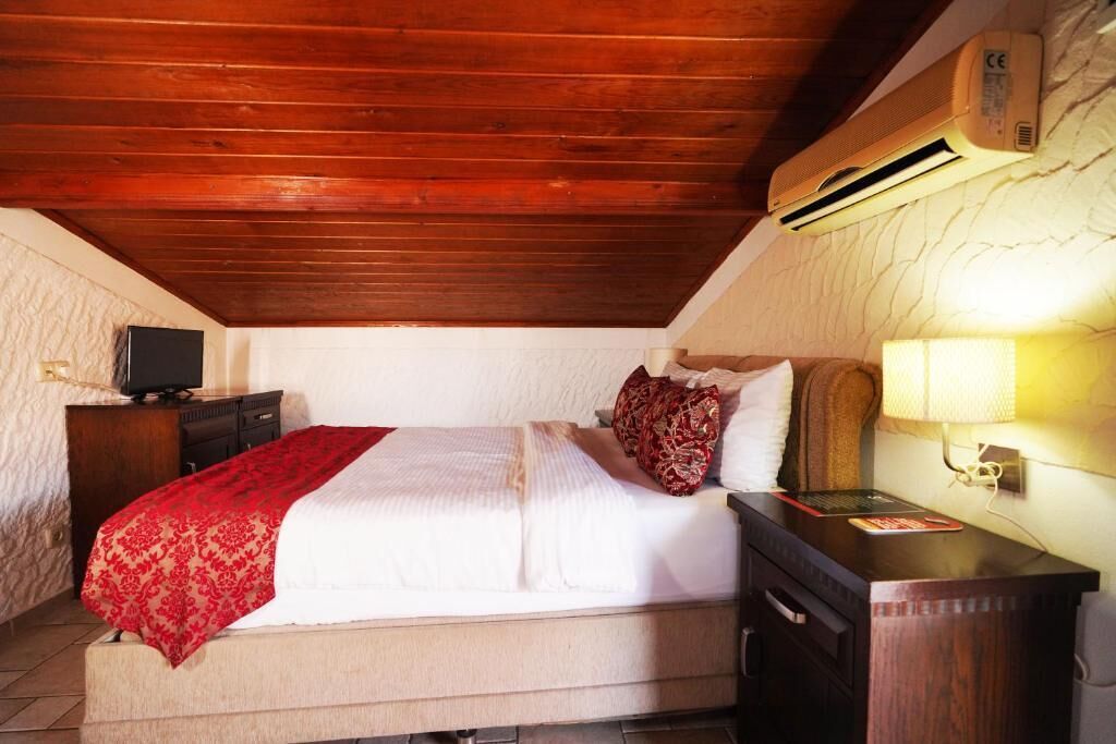 Un dormitorio con techo de madera, una cama con manta roja, una mesita de noche con lámpara y un aire acondicionado.