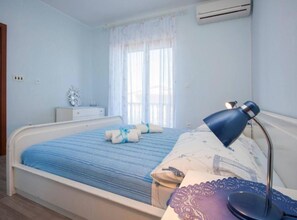 A bedroom with a bed, bedside table, lamp, air conditioning unit, and a window with curtains.
