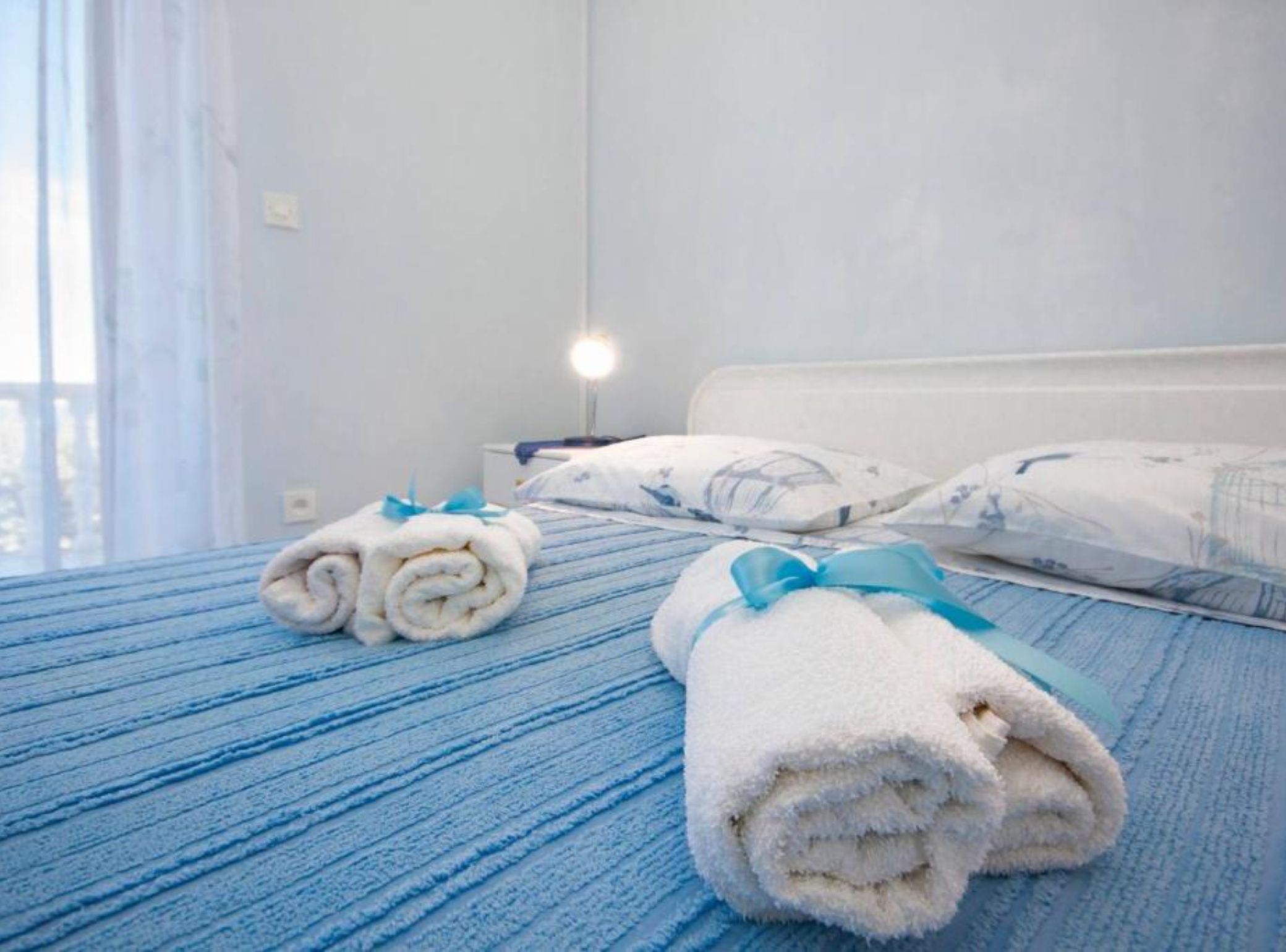 Ein Hotelzimmer mit Bett, zwei zusammengerollte Handtücher mit blauen Bändern, einer Nachttischlampe und einem Fenster mit Vorhängen.