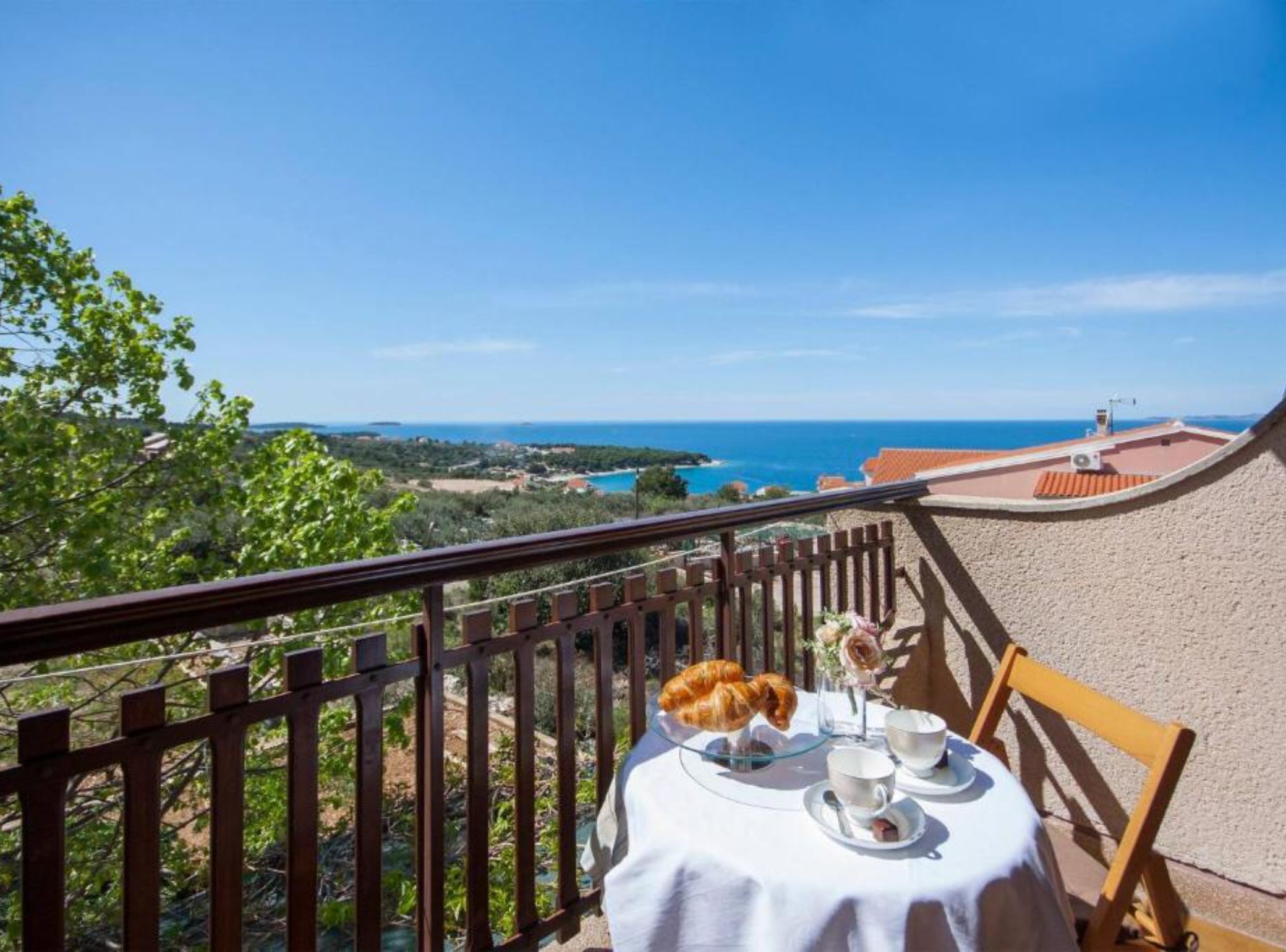 A table with a view of the sea, a balcony with a railing, and a table setting with a croissant and cups.