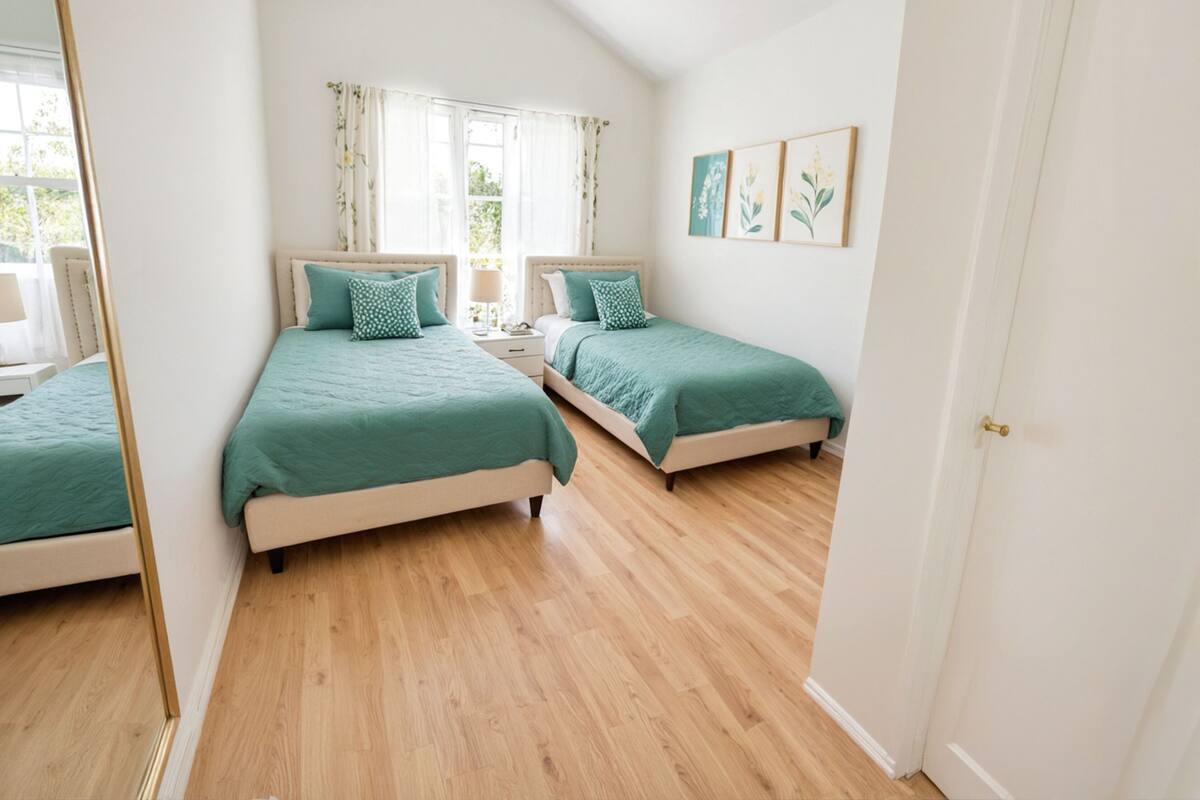 Ein Schlafzimmer mit zwei Betten, einem Spiegel, einem Fenster mit Vorhängen und gerahmten Kunstwerken an der Wand.