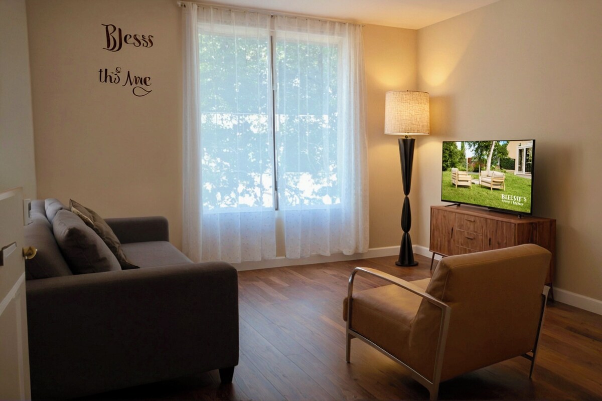 A living room with a sofa, a chair, a TV, a lamp, and a window with sheer curtains.