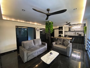 Un salon moderne avec deux canapés gris, une table basse en marbre, un ventilateur de plafond et, au fond, un coin cuisine.