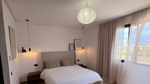 Ein Schlafzimmer mit einem Bett, Nachttischen, einer Pendelleuchte und Blick durch leichte Vorhänge.