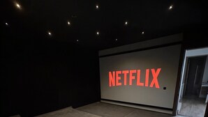 Een donkere kamer met een groot scherm waarop 'NETFLIX' in vetgedrukte letters staat, een paar treden die naar het scherm leiden en een deur aan de rechterkant.