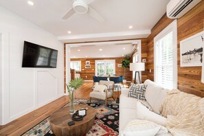 Ein modernes Wohnzimmer mit Flachbildfernseher, Deckenventilator aus Holz, Essbereich mit blauen Stühlen und großem Fenster mit Jalousien.