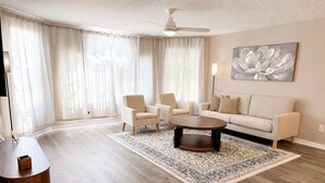 Un salon spacieux avec une grande fenêtre, un ventilateur de plafond et un tableau accroché au mur.