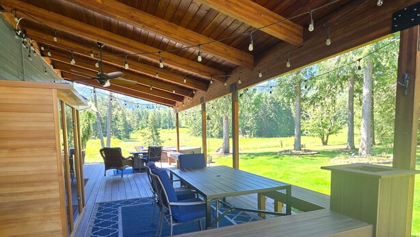 A covered outdoor area with a wooden deck, a table, chairs, and a view of a grassy landscape. - ElkLandia Home & Suite - Sleeps up to 18+ (Sequim)
