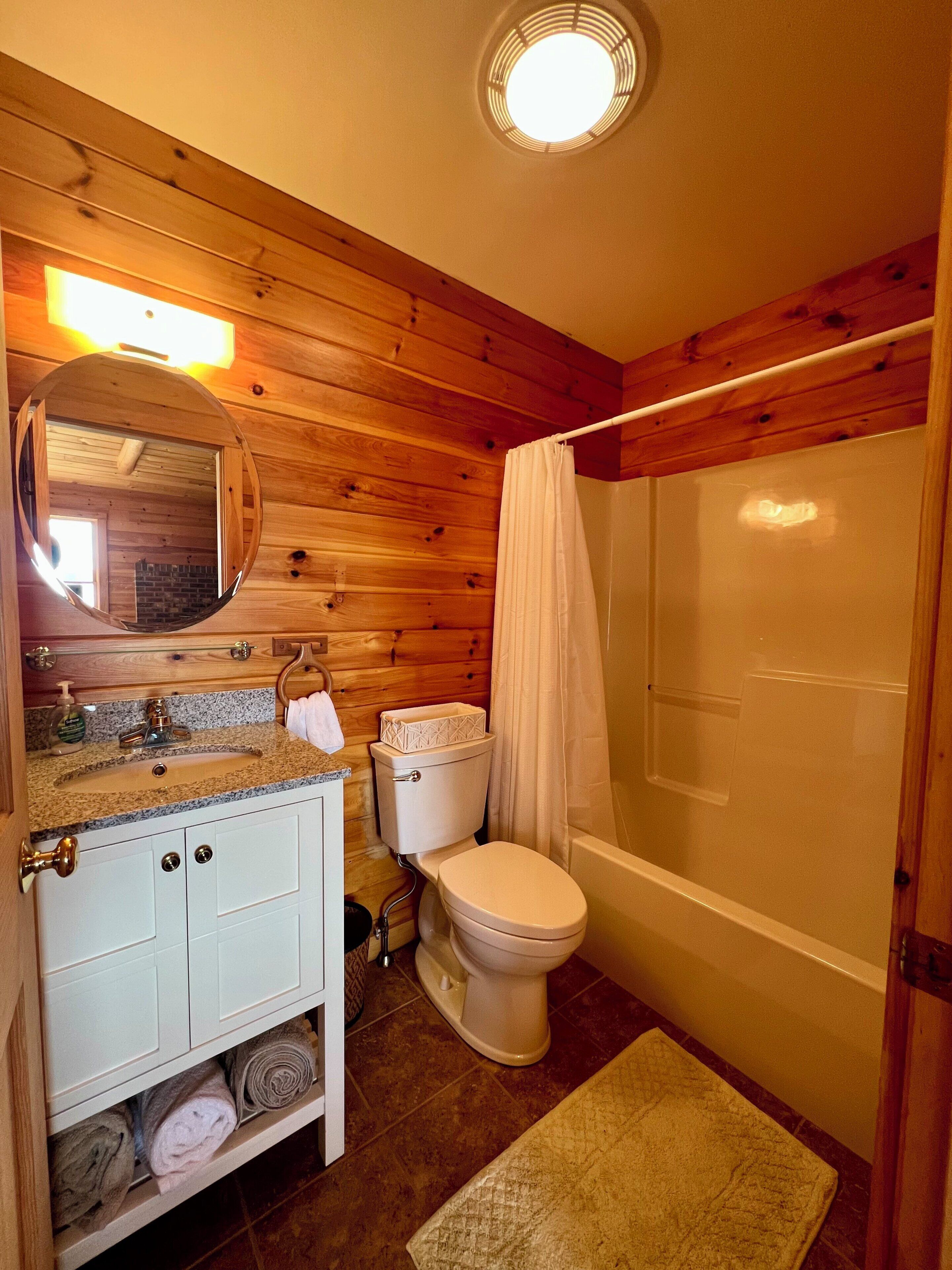Ein Badezimmer mit Holzwänden, einem runden Spiegel, einem Waschbecken, einer Toilette und einer Dusche.