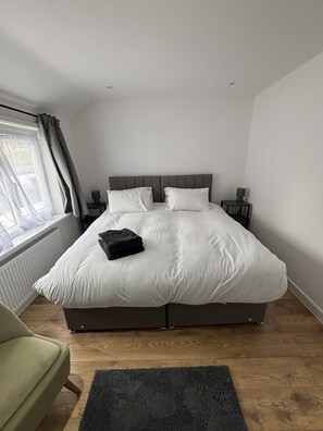 Une chambre à coucher comprenant un lit, une table de chevet, une lampe, une chaise, une fenêtre avec des rideaux et un radiateur.