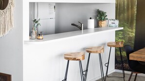 A modern kitchen with white cabinetry, a central island with stools, a macrame wall hanging, and a forest-themed wall. - NEW! Dune and Drift at Crystal Beach 10 (Glen Arbor)