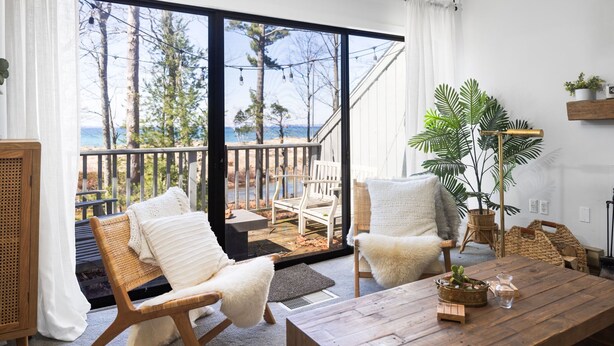 A room with a wooden table, two white chairs, a large potted plant, and a view of the outdoors through a sliding glass door. - NEW! Dune and Drift at Crystal Beach 10 (Glen Arbor)