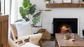 A cozy living room with a fireplace, a wooden coffee table, a wicker chair, and a large plant. - NEW! Dune and Drift at Crystal Beach 10 (Glen Arbor)