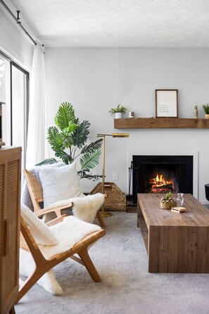 A cozy living room with a fireplace, a wooden coffee table, a wicker chair, and a large plant.