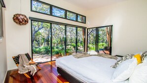 Una camera da letto con un letto grande, vista su una foresta attraverso finestre a tutta parete e pavimento in legno.