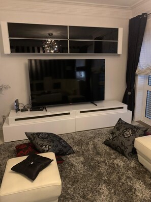 A living room with a flat-screen TV on a white entertainment unit, a sofa, and two patterned cushions.