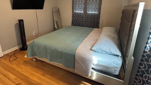 Makuuhuoneessa on sänky, seinälle asennettu televisio, lattiatuuletin ja kuviollinen verho.