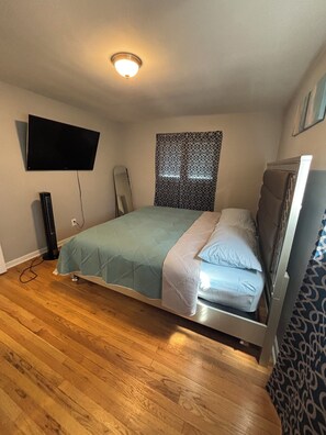 Ein Schlafzimmer mit einem Bett, einem an der Wand montierten Fernseher, einem Standventilator und einem gemusterten Vorhang.